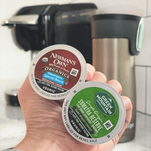 holding two kcup pods with keurig and travel mug in background